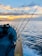Several fishing rods are lined up on the side of a boat as it travels over gentle waves. The sun is setting or rising, casting a warm glow across the sky, which is filled with scattered clouds.