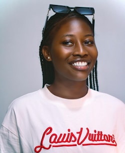 A person smiling confidently at the camera, wearing a white shirt with 'Louis Vuitton' written in red on it. Sunglasses are resting on the person's head, and they have braided hair.