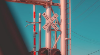 A railroad crossing sign is prominently displayed at an intersection. The sign is mounted on a metal pole along with additional signaling equipment. Overhead, there are several power lines stretching across the sky, which is clear with a bright cyan hue. Part of a railway signaling system, including lights, is visible.