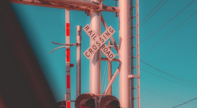 A railroad crossing sign is prominently displayed at an intersection. The sign is mounted on a metal pole along with additional signaling equipment. Overhead, there are several power lines stretching across the sky, which is clear with a bright cyan hue. Part of a railway signaling system, including lights, is visible.