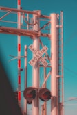 A railroad crossing sign is affixed to a metal pole structure, with various cables and components visible. There are red and white-striped barriers next to the sign. Bright red warning lights are at the base of the crossing sign. The background features a clear, blue sky and some power lines extending horizontally and vertically.