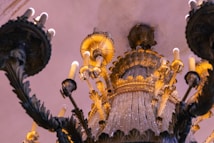 An ornate chandelier with multiple decorative elements including candle-like bulbs, intricate metalwork, and crystal adornments. The design features a blend of dark metal and golden accents, with a luxurious appearance enhanced by soft, warm lighting. The angle of the photo is from below, highlighting the grandeur and elaborate details of the chandelier.