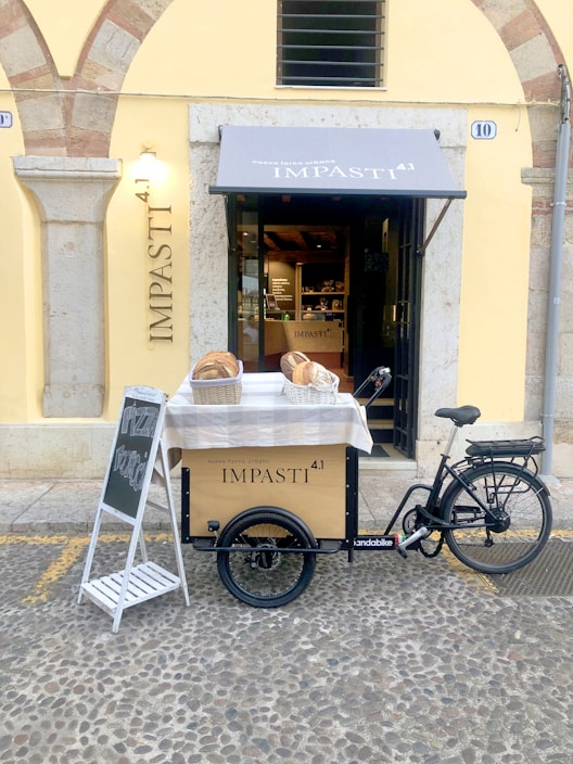 A storefront with a bicycle cart in front, displaying the name 'IMPASTI 4.1'. Two baskets filled with bread are placed on a covered table in front of the shop. A chalkboard sign on a stand beside the cart advertises pizza. The building features stone arches and a light yellow exterior.