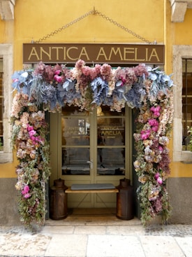 A quaint storefront with a sign reading 'Antica Amelia' adorned with a vibrant floral arrangement. The flowers include a variety of colors such as pink, purple, blue, and green. The door is flanked by two large rustic decorative objects, possibly planters or milk cans. The building is painted in a warm yellow hue.
