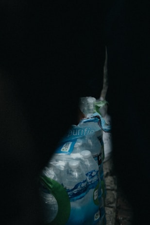 A pack of bottled water is placed on a dimly lit floor. The packaging is partially crumpled, and the bottles are visible inside. The surrounding environment is dark, creating a sense of contrast with the transparency of the plastic bottles.