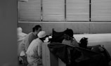Three individuals are gathered near a large cardboard box under an overpass with metal railings in the background. One person is seated, wearing a knit hat, and is looking at the others who are crouching nearby. Objects and bags are scattered around, suggesting a transient or outdoor setting.