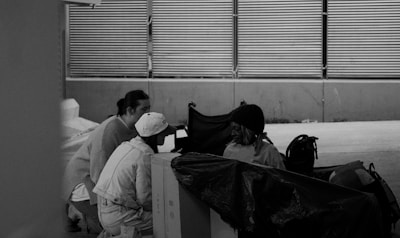Three individuals are gathered near a large cardboard box under an overpass with metal railings in the background. One person is seated, wearing a knit hat, and is looking at the others who are crouching nearby. Objects and bags are scattered around, suggesting a transient or outdoor setting.