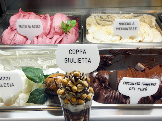 The image depicts a display of various flavors of gelato, each labeled with a sign, in a well-lit ice cream shop. The prominent flavors include 'Frutti di Bosco,' 'Nocciola Piemontese,' 'Cocco Malaysia,' and 'Cioccolato Fondente del Per&uacute;.' In the foreground, there is a dessert cup named 'Coppa Giulietta' topped with nuts and chocolate swirls. Some decorative leaves add a fresh look.