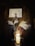 A warmly lit wooden interior with a glowing cylindrical lamp placed on a table. Decorative elements include tall white feathers in a vase, alongside handmade wooden figures resembling reindeer. Above, a painting depicts a blooming tree with white blossoms set against a brick-like background.