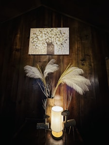 A warmly lit wooden interior with a glowing cylindrical lamp placed on a table. Decorative elements include tall white feathers in a vase, alongside handmade wooden figures resembling reindeer. Above, a painting depicts a blooming tree with white blossoms set against a brick-like background.