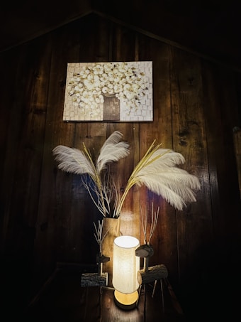 A warmly lit wooden interior with a glowing cylindrical lamp placed on a table. Decorative elements include tall white feathers in a vase, alongside handmade wooden figures resembling reindeer. Above, a painting depicts a blooming tree with white blossoms set against a brick-like background.