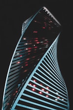 A modern skyscraper with a twisting architectural design stands against a dark background. The building is illuminated by vibrant red and white lights that contrast with its sleek lines. The structure's dynamic curves create a sense of movement and innovation.