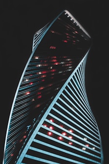 A modern skyscraper with a twisting architectural design stands against a dark background. The building is illuminated by vibrant red and white lights that contrast with its sleek lines. The structure's dynamic curves create a sense of movement and innovation.