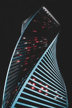 A modern skyscraper with a twisting architectural design stands against a dark background. The building is illuminated by vibrant red and white lights that contrast with its sleek lines. The structure's dynamic curves create a sense of movement and innovation.