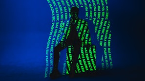A dark silhouette of a person is seated against a deep blue background. Bright green text is projected onto the figure and the backdrop, creating an intricate pattern. The text includes words such as 'ERIC', 'MAGNA', and 'RAJ'. The lighting creates a mysterious and artistic atmosphere.