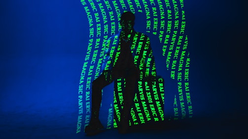 A dark silhouette of a person is seated against a deep blue background. Bright green text is projected onto the figure and the backdrop, creating an intricate pattern. The text includes words such as 'ERIC', 'MAGNA', and 'RAJ'. The lighting creates a mysterious and artistic atmosphere.
