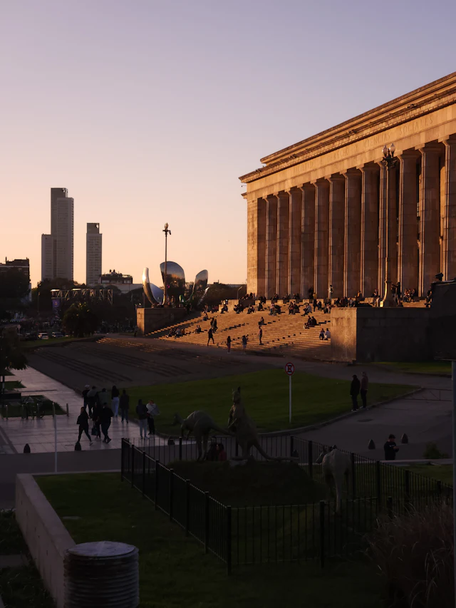 The colonnaded Facultad de Derecho building glowing at dusk with the modern Puerto Madero skyline behind it, seen from Plaza Naciones Unidas in Recoleta.