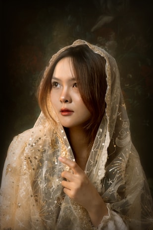A woman is wrapped in an intricately embroidered shawl with gold and silver details, gazing thoughtfully. The background is dark, creating a contrast that highlights her serene expression.