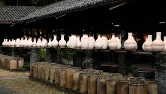 A series of large, unglazed ceramic vases are arranged in a row on a wooden ledge in an outdoor setting. The vases vary slightly in shape and size, showcasing their natural clay texture. The background includes a rustic building with a tiled roof, and the foreground features cylindrical molds or planters.