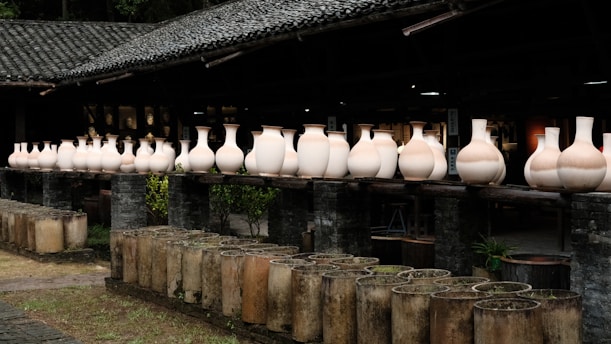 A series of large, unglazed ceramic vases are arranged in a row on a wooden ledge in an outdoor setting. The vases vary slightly in shape and size, showcasing their natural clay texture. The background includes a rustic building with a tiled roof, and the foreground features cylindrical molds or planters.