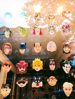 A collection of colorful masks displayed on a window, featuring a variety of characters with different expressions and styles. The top row includes a mix of cartoon and superhero faces, while the lower rows show more distinctive and recognizable figures from various media. The reflection of trees and a building can be seen in the glass.