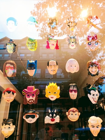 A collection of colorful masks displayed on a window, featuring a variety of characters with different expressions and styles. The top row includes a mix of cartoon and superhero faces, while the lower rows show more distinctive and recognizable figures from various media. The reflection of trees and a building can be seen in the glass.