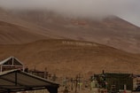 A desolate, foggy desert landscape with a hill adorned with large white letters spelling out 'Poconchile' in multiple places. Several crosses and structures are visible, suggesting a cemetery. The sky is overcast, and the overall terrain is barren and rocky.