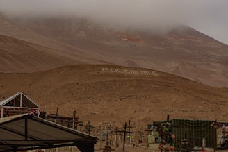 A desolate, foggy desert landscape with a hill adorned with large white letters spelling out 'Poconchile' in multiple places. Several crosses and structures are visible, suggesting a cemetery. The sky is overcast, and the overall terrain is barren and rocky.