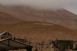 A desolate, foggy desert landscape with a hill adorned with large white letters spelling out 'Poconchile' in multiple places. Several crosses and structures are visible, suggesting a cemetery. The sky is overcast, and the overall terrain is barren and rocky.