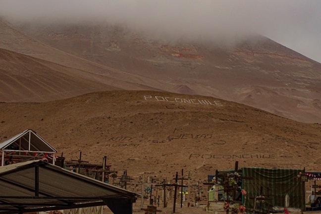 A desolate, foggy desert landscape with a hill adorned with large white letters spelling out 'Poconchile' in multiple places. Several crosses and structures are visible, suggesting a cemetery. The sky is overcast, and the overall terrain is barren and rocky.