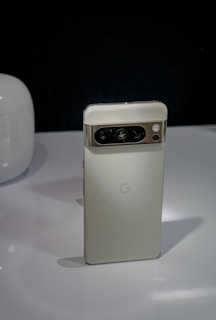 A sleek, vertically-oriented smartphone with a metallic finish on the camera module at the top. The device has a minimalist design with the logo of the brand in the middle of the backside. It is placed on a glossy surface with a dark background, and there's a rounded, white object partially visible on the left.