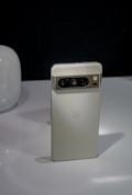 A sleek, vertically-oriented smartphone with a metallic finish on the camera module at the top. The device has a minimalist design with the logo of the brand in the middle of the backside. It is placed on a glossy surface with a dark background, and there's a rounded, white object partially visible on the left.
