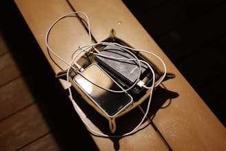 A small basket placed on a wooden surface containing a smartphone, a portable charger, and a tangled white charging cable. The scene is dimly lit, with shadows creating a cozy atmosphere.