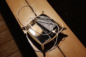 A small basket placed on a wooden surface containing a smartphone, a portable charger, and a tangled white charging cable. The scene is dimly lit, with shadows creating a cozy atmosphere.