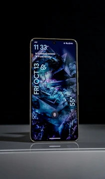 A smartphone is placed upright on a reflective surface against a dark background. The screen displays a digital time of 11:33 with weather information and the date, which reads Friday, October 13. The wallpaper features abstract geometric patterns in dark blue and purple hues, adding a modern and artistic touch.