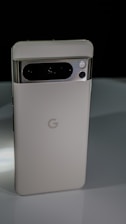 A sleek, modern smartphone with a metallic and glass finish, prominently featuring a camera array on the upper back and the Google logo in the center. The phone is positioned upright against a dark background, highlighting its elegant design.