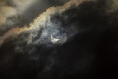 A crescent-shaped solar eclipse is partially obscured by dark, swirling clouds. The scene has an ominous and mysterious atmosphere, with the interplay between the light and shadows creating a dramatic effect.