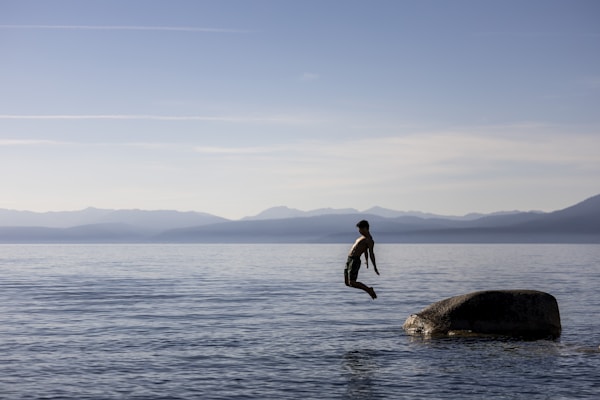 A person is captured mid-air while jumping from a rock into the calm waters of a large body of water, with mountains and a clear sky in the background. The scene conveys a feeling of freedom and adventure.