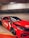 A red sports car with a custom paint job featuring bold graphics is parked in an indoor garage. The vehicle has sleek lines and dark tinted windows, with visible lettering on the side spelling out 'JAWS'. The front of the car displays intricate headlight designs and a glossy finish.