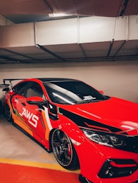 A red sports car with a custom paint job featuring bold graphics is parked in an indoor garage. The vehicle has sleek lines and dark tinted windows, with visible lettering on the side spelling out 'JAWS'. The front of the car displays intricate headlight designs and a glossy finish.