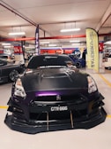 A sleek, modified car with a custom paint job featuring shades of black and purple is prominently displayed in an indoor car show setting. The car's number plate reads 'GTR 666' and it has visible aerodynamic enhancements on the front. The background includes other vehicles, banners, and people, suggesting a bustling environment.