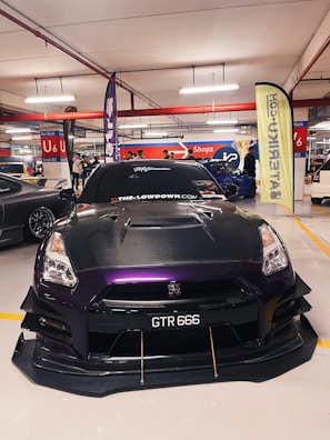 A sleek, modified car with a custom paint job featuring shades of black and purple is prominently displayed in an indoor car show setting. The car's number plate reads 'GTR 666' and it has visible aerodynamic enhancements on the front. The background includes other vehicles, banners, and people, suggesting a bustling environment.