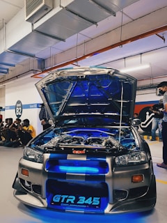 A modified sports car with its hood open, revealing intricate engine components and neon blue lighting. The vehicle is parked in an indoor garage setting with several people gathered around, some sitting on the floor. The license plate reads 'GTR 345'. Overhead, there are industrial pipes and lighting fixtures.