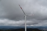 A large wind turbine stands tall against a backdrop of overcast skies with thick, dark clouds. The landscape below features rolling hills and distant mountains, partially shrouded by mist.