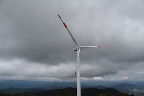 A large wind turbine stands tall against a backdrop of overcast skies with thick, dark clouds. The landscape below features rolling hills and distant mountains, partially shrouded by mist.