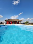 A clear blue swimming pool with a thatched-roof building in the background. The building is painted red and is surrounded by some people and objects, possibly furniture or decor. The sky is bright blue with a few scattered clouds.