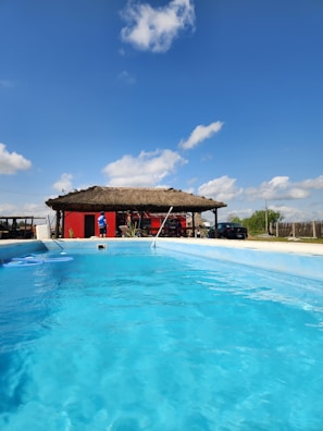 A clear blue swimming pool with a thatched-roof building in the background. The building is painted red and is surrounded by some people and objects, possibly furniture or decor. The sky is bright blue with a few scattered clouds.