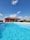 A clear blue swimming pool with a thatched-roof building in the background. The building is painted red and is surrounded by some people and objects, possibly furniture or decor. The sky is bright blue with a few scattered clouds.