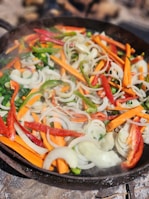 A colorful assortment of sliced vegetables including orange carrots, green and red bell peppers, and white onions are being cooked in a skillet. The vegetables appear fresh and are lightly sautéed, creating a vibrant and appetizing display.