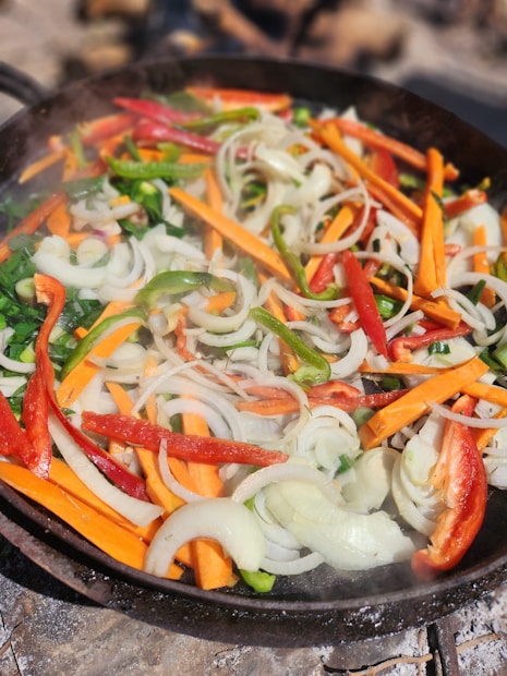 A colorful assortment of sliced vegetables including orange carrots, green and red bell peppers, and white onions are being cooked in a skillet. The vegetables appear fresh and are lightly saut&eacute;ed, creating a vibrant and appetizing display.